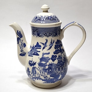 May include: Cream-colored ceramic coffee pot with a blue Willow pattern. The pot features a curved spout, a handle, and a lid with a small knob. The blue pattern depicts a landscape with pagodas, trees, and birds. A classic piece for serving coffee or tea.