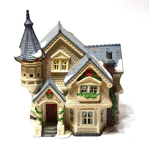 May include: A detailed ceramic Christmas village house with a gray roof, beige walls, and decorative wreaths. The house features a tower, multiple windows, and a chimney, with snow accents on the roof and trim.