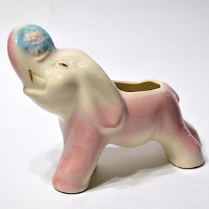 May include: A ceramic elephant figurine in a light pink and cream color scheme. The elephant has a raised trunk and a small opening on its back. The top of the head is light blue with a floral design.