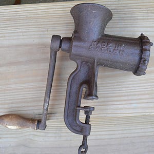 May include: An antique cast iron meat grinder with a wooden handle. The grinder is a dark brown color and has the words "MADE IN SWEDEN" embossed on the side. It features a clamp for securing it to a surface.
