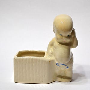May include: A vintage ceramic planter in the shape of a cherubic figure. The off-white planter has a rectangular base with a textured exterior and a small opening. The cherub has blue eyes and a blue accent. A unique decorative item.