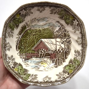 May include: A ceramic bowl featuring a central illustration of a covered bridge in a snowy landscape with green trees. The rim is decorated with a brown and white floral pattern. The bowl has a scalloped edge and a cream-colored background.