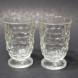 May include: A pair of clear, vintage shot glasses. Each glass features a textured, grid-like design on the body and rests on a small, circular base. The glasses are empty and appear to be made of glass.