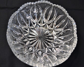 May include: A clear, circular glass bowl featuring an intricate, pressed pattern of radiating starbursts at the center, surrounded by alternating diamond-cut crosshatch textures and smooth, elongated petal-like shapes along the scalloped rim.