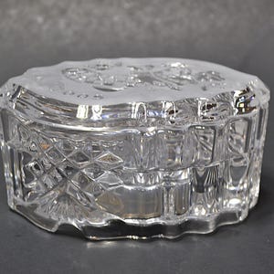 May include: A clear, heart-shaped crystal trinket box with a diamond-cut pattern on the sides. The lid features a frosted design. The box is made of glass and sits on a dark surface. A decorative storage item.