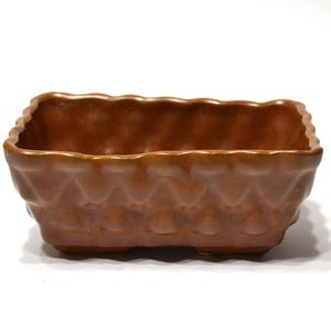 May include: A rectangular ceramic planter with a warm brown glaze. The exterior features a repeating embossed geometric pattern of rounded shapes along the sides, and the top rim has a scalloped, wavy edge design throughout the entire perimeter.