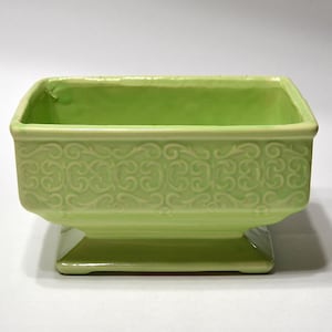May include: A rectangular light green ceramic planter with a raised scrollwork pattern along the exterior sides. The vessel features a wide opening, a tapered base, and a smooth, glossy finish that reflects light across its surface.