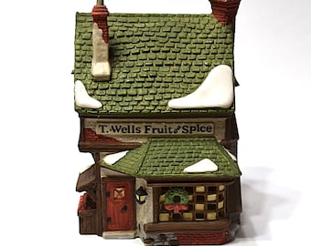 Può includere: Un negozio in miniatura in ceramica con un tetto di tegole verdi e un camino in mattoni rossi. Il negozio ha una porta rossa e un'insegna che recita "T. Wells Fruit & Spice". Una ghirlanda natalizia è appesa sopra la porta.