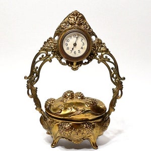 May include: A gold-toned, ornate, decorative trinket box with a hinged lid and a small clock on top. The box is decorated with floral and scrollwork designs. The clock face has black hands and Roman numeral markings.