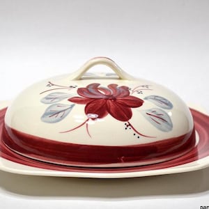 May include: Cream-colored ceramic butter dish with a red floral design on the lid. The lid has a handle and sits on a matching plate with a red border. The dish is a decorative kitchenware item.