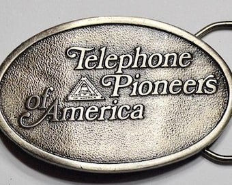 May include: Oval-shaped, silver-toned metal belt buckle. The buckle features the words "Telephone Pioneers of America" in a decorative font. The buckle has a textured surface and a simple metal loop for the belt.