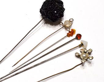 May include: A collection of vintage hat pins with decorative tops. The pins feature various designs, including a black textured ball, a heart-shaped stone, amber-colored beads, and a pearl and rhinestone flower. The pins are arranged on a white surface.