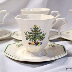 May include: Set of off-white teacups and saucers with a Christmas tree design. The cups have a unique handle shape and sit on matching saucers with a green trim. The Christmas tree is decorated with ornaments and gifts.