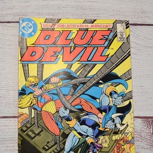 May include: A vintage comic book titled "Blue Devil" with a dynamic illustration of two costumed figures in action. The cover features bold yellow and blue text, with the DC Comics logo. The comic is from January 1985.