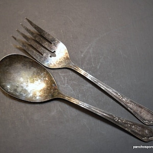 May include: A vintage silver spoon and fork set. The spoon has a rounded bowl, and the fork has four tines. Both utensils have decorative handles. The set is displayed on a dark gray surface.