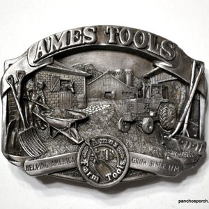 May include: A rectangular silver-toned metal belt buckle featuring a relief scene of a farm with a barn, a tractor, and a person using a wheelbarrow. The text AMES TOOLS is at the top, and HELPING AMERICA GROW SINCE 1774 is at the bottom.