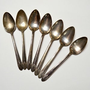 May include: A set of six vintage silver spoons arranged in a fan shape. The spoons have oval bowls and ornate detailing on the handles. The spoons are made of a shiny, reflective metal.