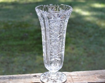 May include: A clear glass vase featuring a flared, serrated rim and a body adorned with intricate, geometric starburst and cross-hatch patterns. The vase stands on a circular, faceted pedestal base, reflecting light through its cut glass design.