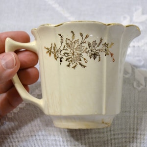 May include: A cream-coloured vintage creamer with a gold floral design. The creamer has a scalloped rim and a handle. The floral design is in shades of gold and brown. The creamer is on a white surface.