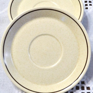 May include: Two round ceramic saucers with a speckled cream-colored finish and a single dark brown circular line accent near the outer rim. The saucers feature a central indentation designed to hold a cup, with a smooth, glossy surface texture.
