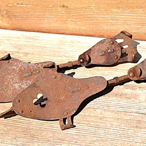 May include: Two antique metal ice skates with a heavy rusted finish. Each skate features a teardrop-shaped plate with a clover-shaped cutout, a long metal runner, and a mechanical heel adjustment mechanism with a series of small rectangular slots.