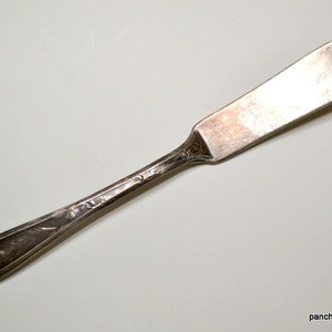 May include: A vintage silver butter knife with a rounded blade and ornate handle. The knife has a slightly tarnished appearance, suggesting age and use. The handle features delicate embossed detailing.