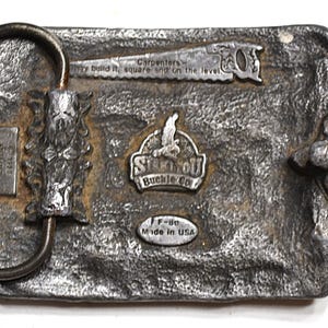 May include: A rectangular, silver-toned metal belt buckle with a textured surface. It features a saw, the text "Carpenters - build it, square and on the level," and the "Stin-OU Buckle Co" logo. The buckle is marked "Made in USA."