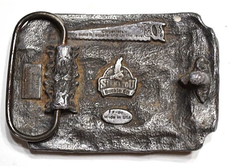 May include: A rectangular, silver-toned metal belt buckle with a textured surface. It features a saw, the text "Carpenters - build it, square and on the level," and the "Stin-OU Buckle Co" logo. The buckle is marked "Made in USA."