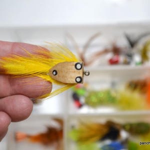 May include: A handmade fishing lure with a wooden body and bright yellow feathers. The lure has black eye details and is held in a hand. Other fishing lures are visible in the background.