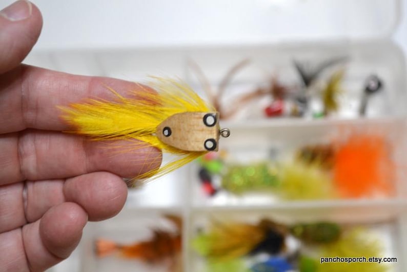 May include: A handmade fishing lure with a wooden body and bright yellow feathers. The lure has black eye details and is held in a hand. Other fishing lures are visible in the background.