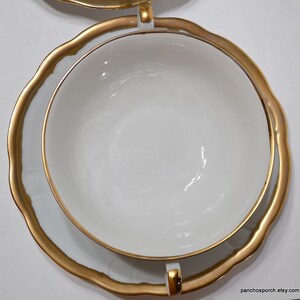 May include: Overhead view of a white teacup and saucer set with gold trim. The cup sits inside the saucer, both with a scalloped edge. The gold trim is thick and ornate, adding a touch of elegance to the classic design. The set is clean and bright.