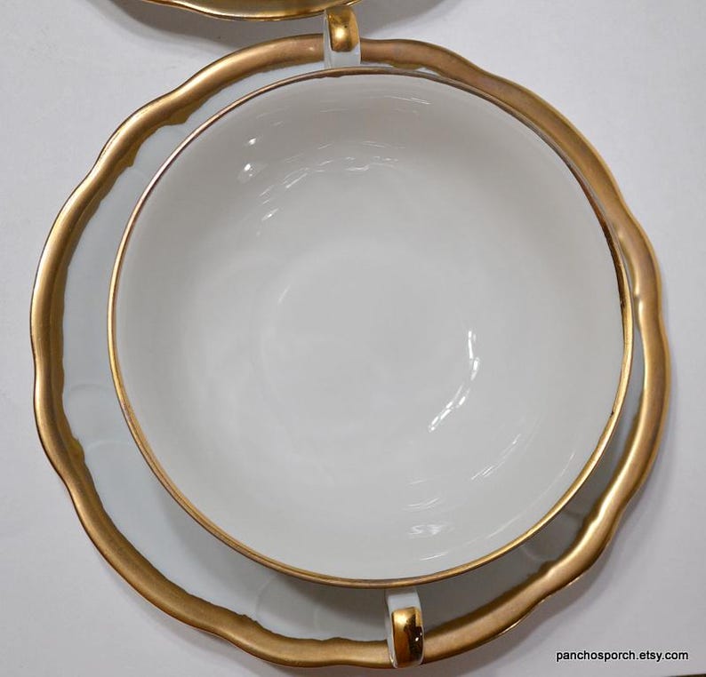 May include: Overhead view of a white teacup and saucer set with gold trim. The cup sits inside the saucer, both with a scalloped edge. The gold trim is thick and ornate, adding a touch of elegance to the classic design. The set is clean and bright.