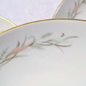May include: Close-up of several white porcelain plates with a gold rim. Each plate features a delicate design of wheat stalks in shades of pink, green, and gray. The plates are arranged to show the repeating pattern.