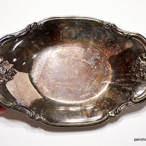 May include: An antique silver-plated serving dish with ornate floral and scroll detailing. The oval dish has a tarnished surface and a decorative rim. The dish is held by a hand against a white background. The Etsy shop name is visible.