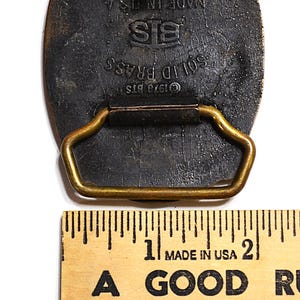 May include: Close-up of a vintage solid brass belt buckle. The buckle is dark with a gold-colored loop and a rectangular shape. The text "SOLID BRASS", "&copy;1978 BTS", and "MADE IN USA" are visible.