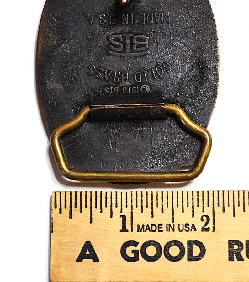 May include: Close-up of a vintage solid brass belt buckle. The buckle is dark with a gold-colored loop and a rectangular shape. The text "SOLID BRASS", "&copy;1978 BTS", and "MADE IN USA" are visible.