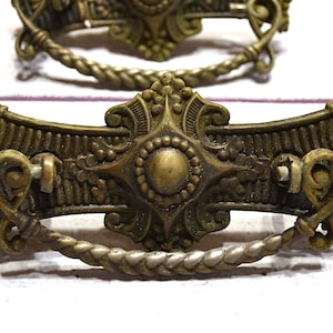 May include: Two antique brass drawer pulls with ornate detailing. Each pull features a decorative central medallion, intricate scrollwork, and a twisted rope-style handle. The hardware is aged with a vintage patina.