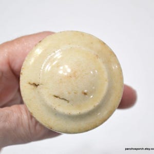 May include: A round, off-white ceramic knob with a slightly domed top. The knob has a circular design with a raised center and a subtle, aged appearance. The surface shows minor imperfections and a light brown stain.