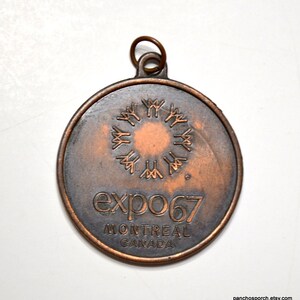 May include: A round, bronze-colored Expo 67 Montreal Canada commemorative medal. The medal features a central design of stylized figures around a circle, with the text "expo 67" and "MONTREAL CANADA" below. A small loop is at the top.