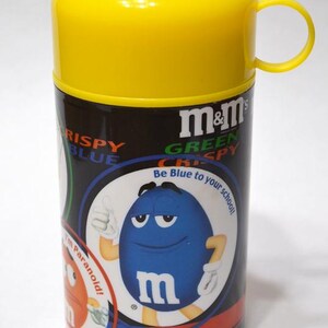 May include: A yellow and black M&M's Crispy candy container with a yellow lid and handle. The container features a blue M&M's character with the text "Be Blue to your school!" and the M&M's logo.