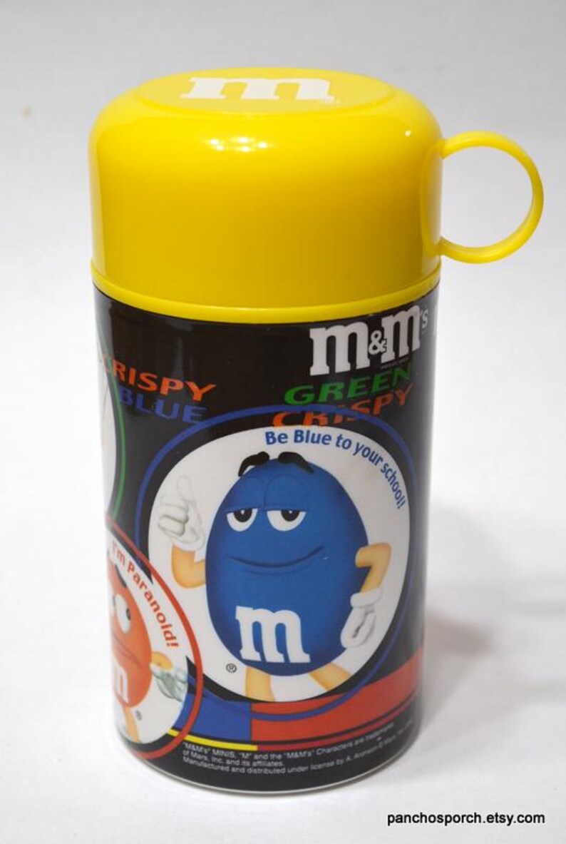 May include: A yellow and black M&M's Crispy candy container with a yellow lid and handle. The container features a blue M&M's character with the text "Be Blue to your school!" and the M&M's logo.