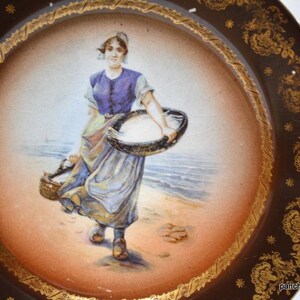 Op de afbeelding: Antieke decoratieve schotel met een handgeschilderde sc&egrave;ne van een vrouw die manden op een strand draagt. De schotel heeft een bruine rand met gouden bloemen- en sterontwerpen en een cr&egrave;mekleurig midden.