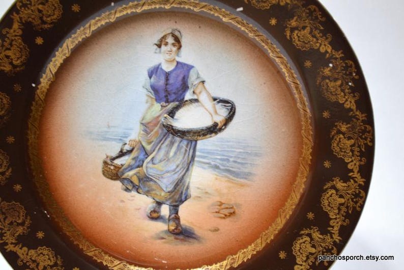 Op de afbeelding: Antieke decoratieve schotel met een handgeschilderde sc&egrave;ne van een vrouw die manden op een strand draagt. De schotel heeft een bruine rand met gouden bloemen- en sterontwerpen en een cr&egrave;mekleurig midden.
