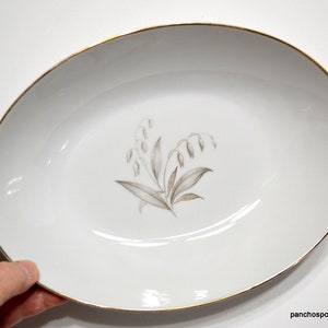 May include: White oval serving dish with a gold rim and a floral design in the center. The floral design features a cluster of wheat stalks in shades of gray and brown. The dish is held by a hand.