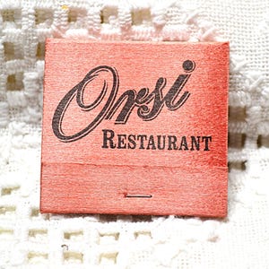May include: A vintage matchbook with a salmon-colored cover. The matchbook features the name "Orsi" in a stylized script above the word "Restaurant" in a serif font. A small, silver staple is visible at the bottom.