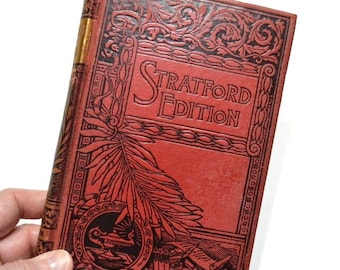 以下が含まれることがあります： 「Stratford Edition」のタイトルがエンボス加工された赤い表紙のヴィンテージ本。表紙には複雑な黒いイラストと金色の背表紙があります。本は人の手で持たれています。
