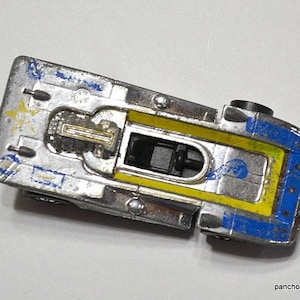 May include: Vintage die-cast metal toy car with a silver body, yellow and blue accents. The toy car has a black interior and wheels. The car shows signs of wear and tear, with paint chips and scratches.