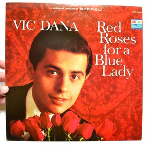 May include: Album cover for Vic Dana's "Red Roses for a Blue Lady." The cover features a portrait of Vic Dana with red roses at the bottom and the album title in white text. The background is a textured red.