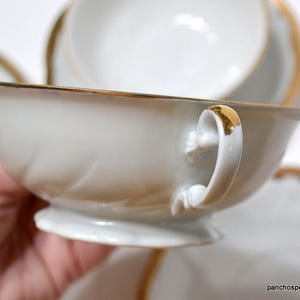 May include: White teacup with a gold rim and handle accents. The teacup is part of a set, with a matching saucer visible in the background. The handle and rim have a gold finish, adding a touch of elegance to the porcelain.