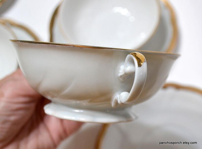 May include: White teacup with a gold rim and handle accents. The teacup is part of a set, with a matching saucer visible in the background. The handle and rim have a gold finish, adding a touch of elegance to the porcelain.